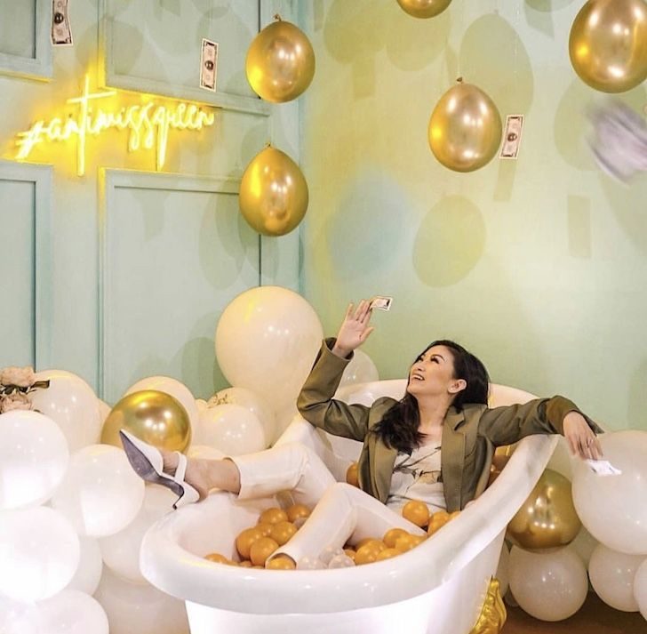 A visitor taking a selfie in a bathtub of orange balls under gold balloons at Baluun by HALUU