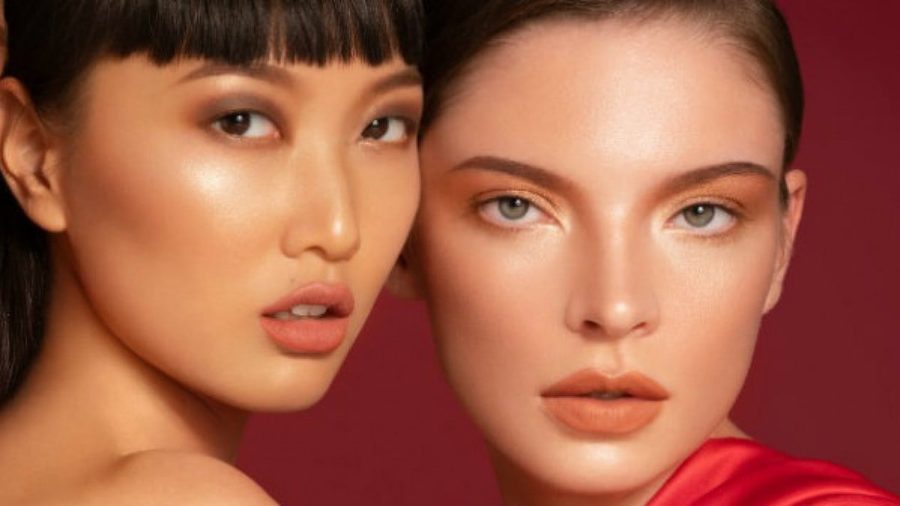 Two models with glowing skin and coral lips posed against a deep red backdrop